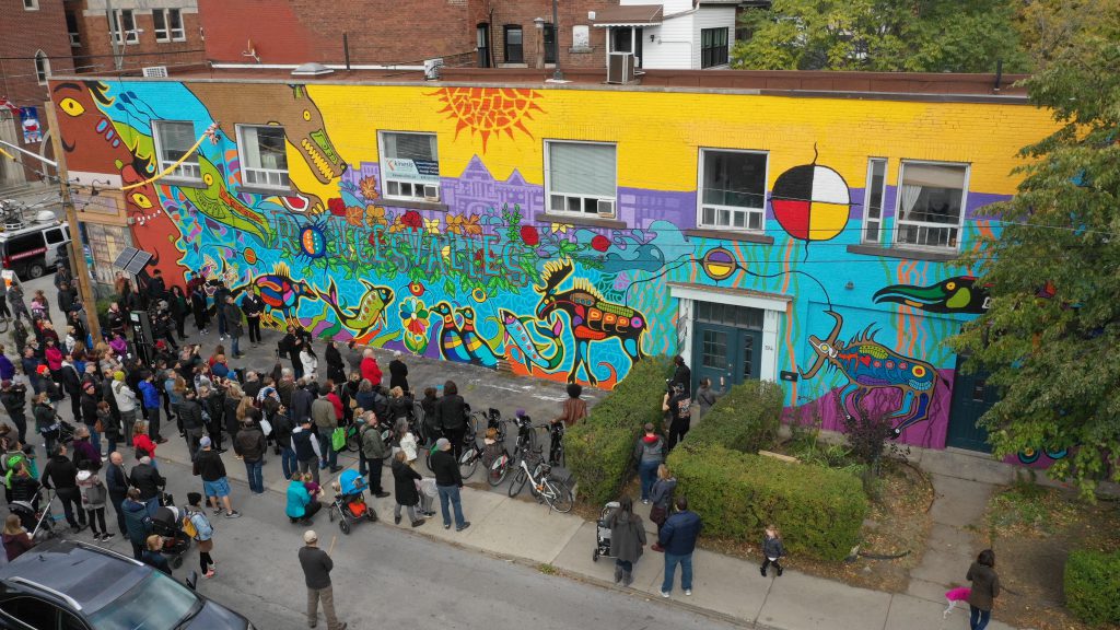 Crowd looking over the mural which features Woodlands style animals and symbols by Philip Cote and the word Roncesvalles intertiwned with foliage by Jim Bravo